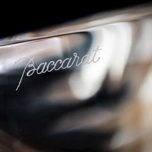 Puede incluir: Un objeto de vidrio transparente y liso que presenta la palabra Baccarat grabada en cursiva. La superficie es transl&uacute;cida con sutiles reflejos de luz y contornos curvos, mostrando el nombre de la marca grabado en el material.