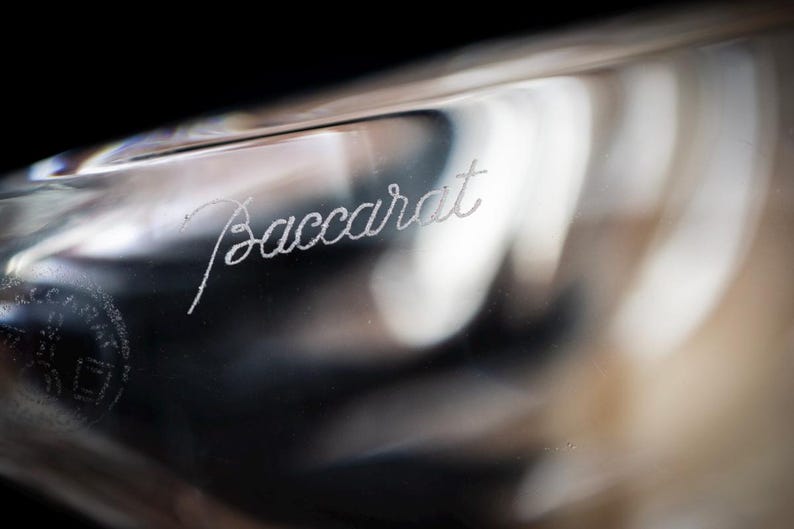 Puede incluir: Un objeto de vidrio transparente y liso que presenta la palabra Baccarat grabada en cursiva. La superficie es transl&uacute;cida con sutiles reflejos de luz y contornos curvos, mostrando el nombre de la marca grabado en el material.