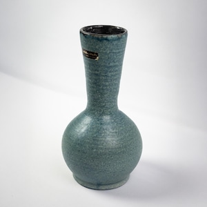 以下が含まれることがあります： 丸みを帯びたベースと細い首を持つ、ターコイズ色のセラミック花瓶。花瓶はテクスチャ加工された表面と濃い茶色の内側を持っています。小さなラベルが首に貼られています。花瓶はおそらく装飾目的で使用されます。