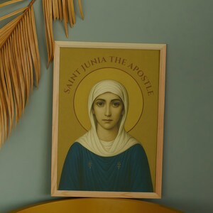 May include: A framed print of Saint Junia the Apostle, featuring a portrait of a woman with a halo, wearing a white head covering and blue robe. The text "SAINT JUNIA THE APOSTLE" is in a circular pattern around the halo. A dried palm leaf is in the background.