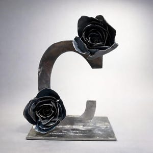 Puede incluir: Escultura metálica con dos rosas negras. Las rosas están unidas a una estructura metálica curva sobre una base rectangular. La escultura tiene una estética oscura e industrial.