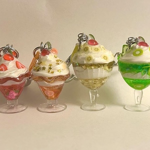 Puede incluir: Cuatro llaveros en miniatura con forma de postres, representando parfaits. Cada parfait tiene una cobertura de fruta diferente, incluyendo fresas, naranjas, plátanos y kiwi. Los llaveros están en copas transparentes con aros plateados.