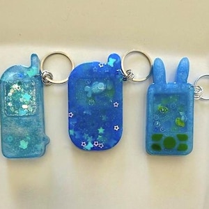Könnte beinhalten: Drei blaue Schlüsselanhänger aus Kunstharz in Form von Retro-Handys. Die Schlüsselanhänger haben silberne Schlüsselringe und sind mit Glitzer und kleinen dekorativen Elementen gefüllt. Einer hat ein Hasenohren-Design.