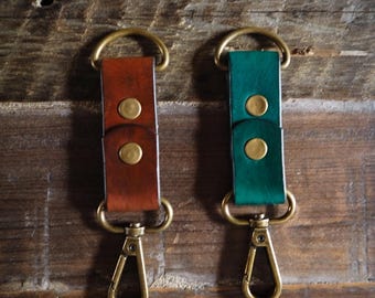 May include: Two leather keychains, one brown and one green, with brass hardware. Each keychain features a leather strap secured with two brass rivets and a brass clasp. The keychains are displayed on a wooden surface.