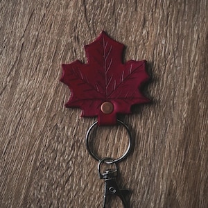 May include: A burgundy leather keychain in the shape of a maple leaf. The leaf has detailed veining and a copper rivet. It is attached to a silver keyring and a lobster clasp.