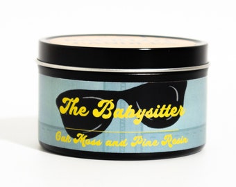 May include: A black tin candle with a blue and white label. The label features a pair of sunglasses and the text "The Babysitter Oak Moss and Pine Resin".