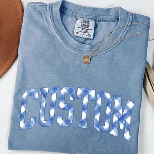 May include: A light blue Comfort Colors t-shirt with the word "CUSTOM" in a blue and white gingham pattern. A gold necklace with a pendant rests on the shirt. A brown hat and dried plant are in the background.