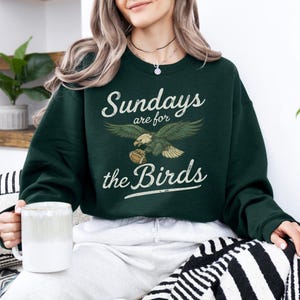 A woman wearing a green sweatshirt with the text "Sunday's are for the Birds" is sitting on a couch, holding a mug in her hand.