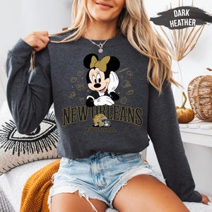 A woman wearing a grey sweatshirt with a Minnie Mouse graphic and text that says "New Orleans Football" is sitting in a room with a pumpkin decoration.