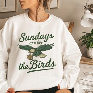 A woman wearing a white sweatshirt with a graphic design that says "Sunday are for the Birds" and features an eagle holding a football.