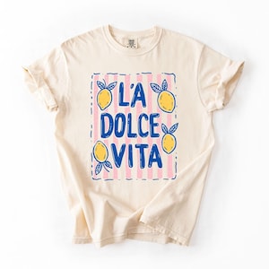 A white t-shirt with a graphic design featuring the text "LA Dolce Vita" surrounded by lemon and lime illustrations.