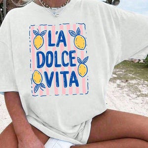 A person wearing a white t-shirt with a graphic design featuring the text "L'A Dolce Vita" and illustrations of lemons.