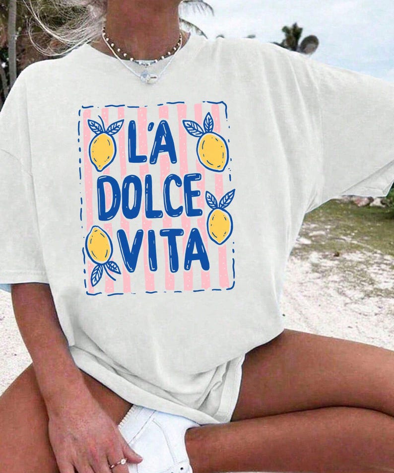 A person wearing a white t-shirt with a graphic design featuring the text "L'A Dolce Vita" and illustrations of lemons.