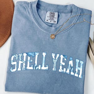May include: A light blue Comfort Colors t-shirt with the words "SHELL YEAH" in white block letters with a blue floral pattern. A gold necklace with a round pendant rests on the shirt. A brown hat and dried leaves are in the background.