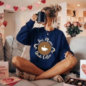 May include: Navy blue sweatshirt with the text "This Mama Needs Coffee" in a circular design. The design features a coffee cup and steam. The person is holding a black mug.