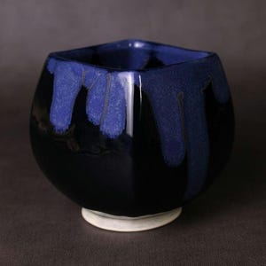 May include: A handmade ceramic bowl with a square top and rounded body. The bowl is glazed in a glossy black with blue drips cascading from the rim. It sits on a small, white circular base, showcasing a unique, artistic design.