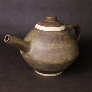 May include: A handcrafted ceramic teapot with a rounded body and a lid. The teapot is primarily olive green with a white band around the lid and base. It has a curved handle and a spout.