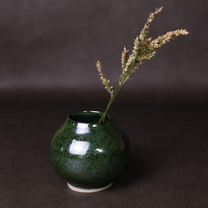 May include: A small, round, dark green ceramic vase with a glossy finish. The vase has a white base and contains a sprig of dried, light-colored flowers. The vase is displayed against a dark background.