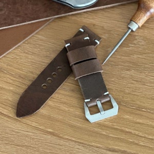 May include: A brown leather watch strap with white stitching and a silver buckle. The strap is shown on a wooden surface.