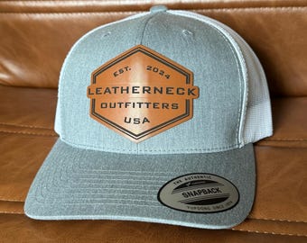 Könnte beinhalten: Eine graue und weiße Baseballkappe mit einem braunen Lederpatch, auf dem "EST. 2024 LEATHERNECK OUTFITTERS USA" steht. Die Kappe hat ein weißes Netzrücken und ein schwarzes Etikett "The Authentic Yupoong Snapback Yupoong Since 1974".