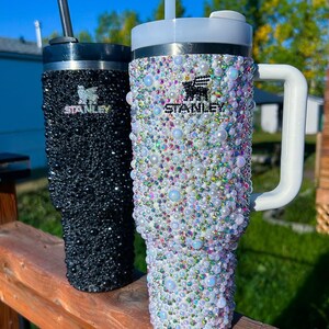 Puede incluir: Dos vasos Stanley con pedrer&iacute;a. Uno es negro con tapa y pajita negras, el otro es blanco con asa y tapa blancas. Ambos vasos est&aacute;n cubiertos de gemas y perlas iridiscentes. El logotipo de Stanley es visible en ambos.