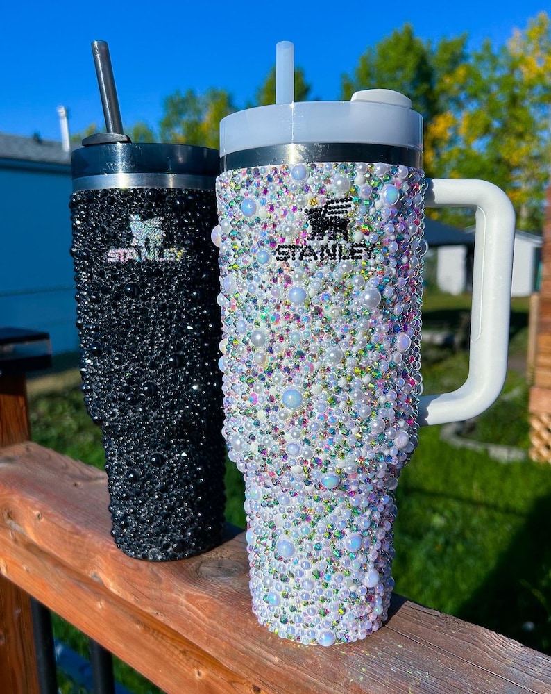 Puede incluir: Dos vasos Stanley con pedrer&iacute;a. Uno es negro con tapa y pajita negras, el otro es blanco con asa y tapa blancas. Ambos vasos est&aacute;n cubiertos de gemas y perlas iridiscentes. El logotipo de Stanley es visible en ambos.