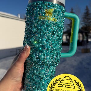 Puede incluir: Un vaso de color turquesa cubierto de cuentas iridiscentes, con un asa y tapa a juego. El vaso tiene una pajita y un logotipo amarillo que dice "THE GILDED HIVE BESPOKE GIFTS".