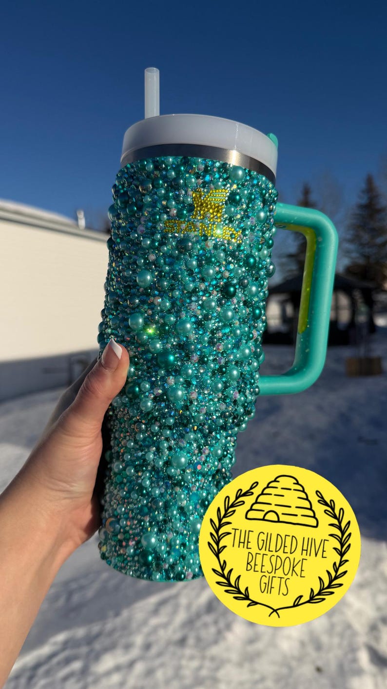 Puede incluir: Un vaso de color turquesa cubierto de cuentas iridiscentes, con un asa y tapa a juego. El vaso tiene una pajita y un logotipo amarillo que dice "THE GILDED HIVE BESPOKE GIFTS".