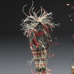 May include: Three small figurative sculptures standing on circular wooden log bases. Each figure features a dark, textured body wrapped in woven fibers of red, white, green, and brown, with wild, fibrous hair and small beads attached to the strands.