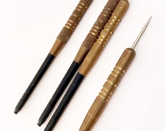 May include: Four vintage darts with brass and black accents. The darts have a metal tip and a textured grip. The barrels are a mix of brass and bronze, with black shafts. The darts are arranged diagonally against a white background.