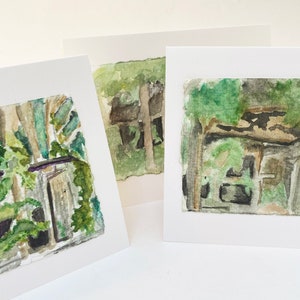 May include: Three watercolor paintings of rustic buildings, each framed in white. The artwork features muted greens, browns, and grays, depicting overgrown structures. The paintings are on textured paper, suggesting a handmade quality.
