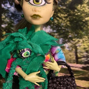 May include: A green-skinned doll with one large green eye and a green fabric doll with one large green eye. The doll is wearing a green and purple shirt and a black purse with a swirl design.