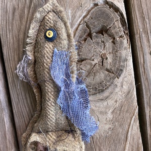 May include: A handmade, rustic-looking, brown and blue fabric doll with a yellow button for an eye. The doll is hanging from a string on a wooden surface.