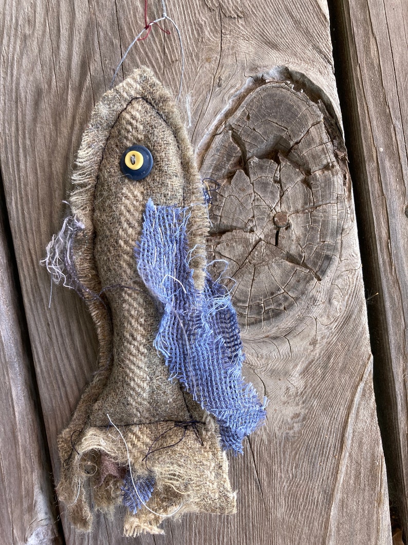 May include: A handmade, rustic-looking, brown and blue fabric doll with a yellow button for an eye. The doll is hanging from a string on a wooden surface.