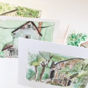 May include: Watercolor paintings of rustic houses and trees, mounted on white cardstock. The artwork features muted greens, browns, and grays, capturing a serene, rural landscape. The paintings are displayed at various angles, showcasing the artist's style.