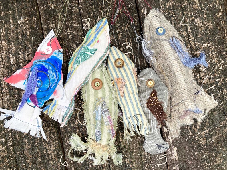 May include: Five handmade fabric fish ornaments in various colors and patterns, including blue, red, white, green, and brown. The fish are hanging from a wooden surface.