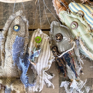 May include: Five handmade fabric fish ornaments hanging from a branch. The fish are made from different fabrics and have different colors and patterns. Each fish has a button for an eye.