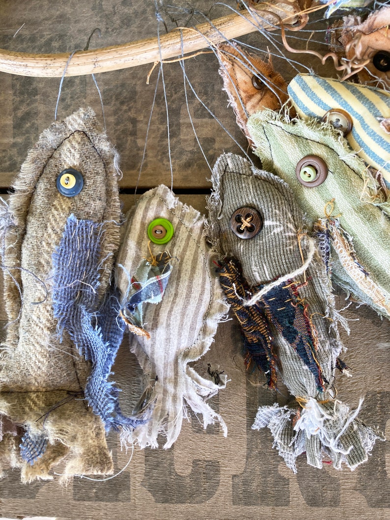 May include: Five handmade fabric fish ornaments hanging from a branch. The fish are made from different fabrics and have different colors and patterns. Each fish has a button for an eye.
