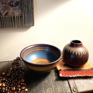 May include: A ceramic bowl with a dark brown exterior, a bright blue interior band, and a central orange circle with green accents. Beside it sits a small, round, dark brown ceramic vase featuring a cream-colored band with a repeating brown pattern.