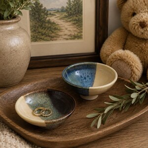 May include: Two small ceramic bowls with a two-tone glaze finish, one featuring blue and cream colors and the other green and cream, resting on a large oval wooden tray. Two gold-toned metal rings are placed inside the green and cream bowl.