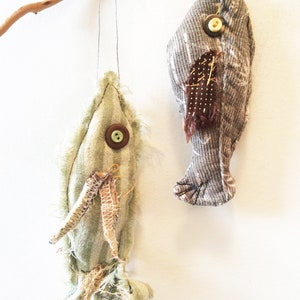 May include: Two handmade fabric fish ornaments hanging from a branch. The fish are made from different fabrics and have buttons for eyes. One fish is green and the other is gray.