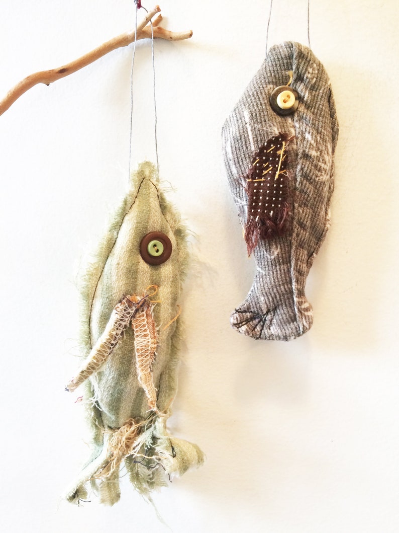 May include: Two handmade fabric fish ornaments hanging from a branch. The fish are made from different fabrics and have buttons for eyes. One fish is green and the other is gray.