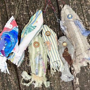 May include: Five handmade fabric fish ornaments in various colors and patterns, including blue, red, white, green, and brown. The fish are hanging from a wooden surface.