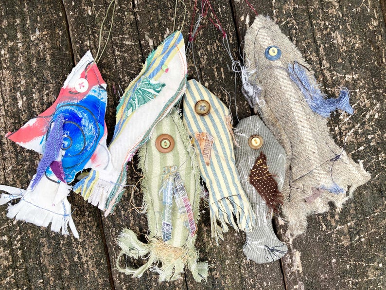 May include: Five handmade fabric fish ornaments in various colors and patterns, including blue, red, white, green, and brown. The fish are hanging from a wooden surface.