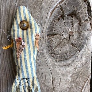 May include: A handmade yellow and blue striped fish toy with a button eye and frayed edges. The fish is hanging on a weathered wooden surface.