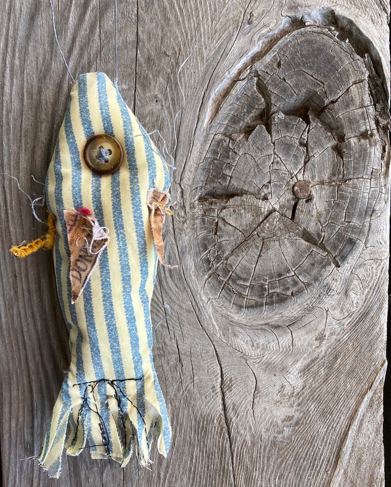 May include: A handmade yellow and blue striped fish toy with a button eye and frayed edges. The fish is hanging on a weathered wooden surface.