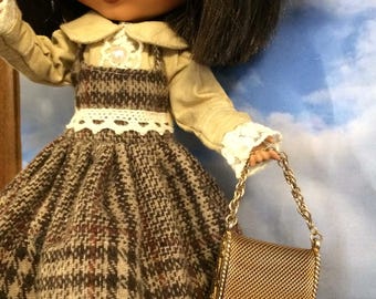 May include: A doll wearing a brown and white plaid dress with a white lace trim and a brown leather belt. The doll is holding a gold mesh purse with a chain strap.