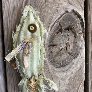 May include: A green fabric fish ornament with a brown button and frayed edges. The fish is hanging from a string on a wooden background.