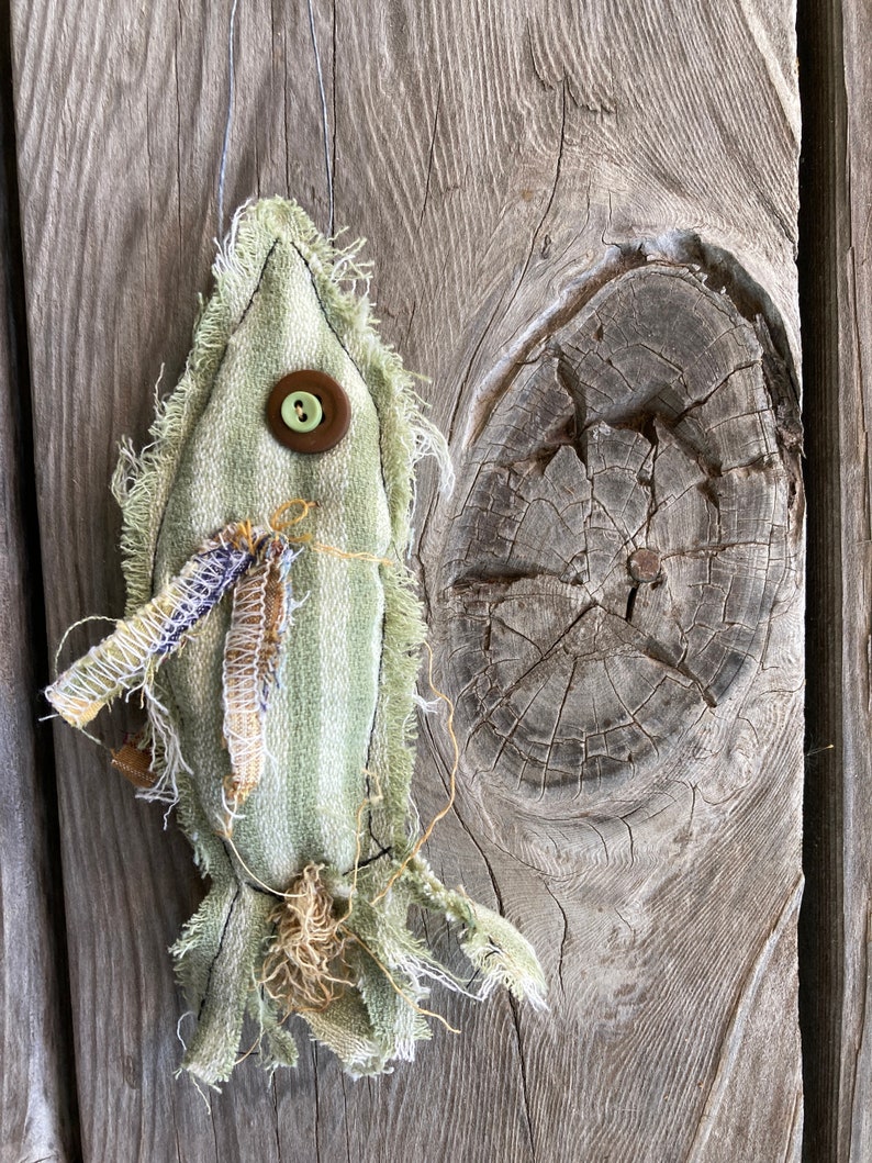 May include: A green fabric fish ornament with a brown button and frayed edges. The fish is hanging from a string on a wooden background.