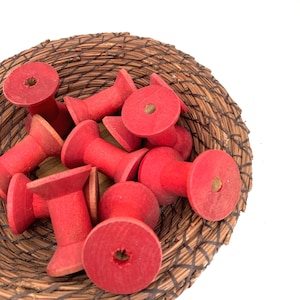 May include: A collection of small, red wooden spools inside a woven basket. The spools have a cylindrical shape with a wider top and bottom, and a hole through the center. The basket is made of brown, natural fibers.